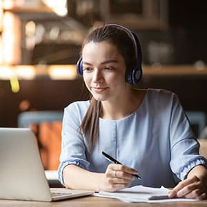 Focused woman wearing headphones using laptop, writing notes